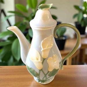 May include: A ceramic teapot with a white and pale blue body, adorned with yellow calla lily designs and green accents. The handle and spout are curved, and the lid is topped with a green leaf. The teapot is displayed on a wooden surface.