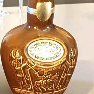 K&ouml;nnte beinhalten: Eine braune Keramikflasche Royal Salute Scotch Whisky. Die Flasche hat ein silbernes Etikett und ein gepr&auml;gtes Design mit Figuren und dem Markennamen. Die Flasche steht auf einer hellen Oberfl&auml;che.