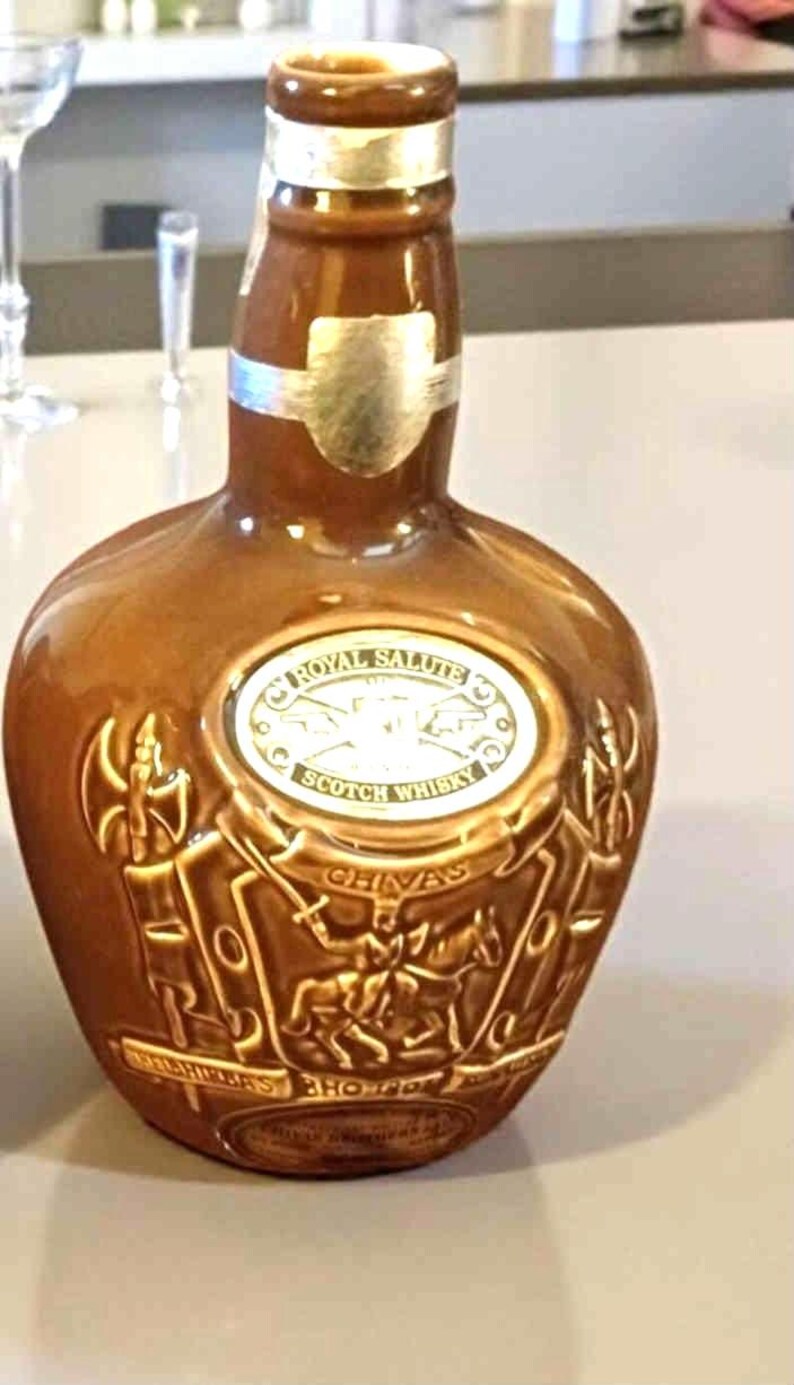 K&ouml;nnte beinhalten: Eine braune Keramikflasche Royal Salute Scotch Whisky. Die Flasche hat ein silbernes Etikett und ein gepr&auml;gtes Design mit Figuren und dem Markennamen. Die Flasche steht auf einer hellen Oberfl&auml;che.