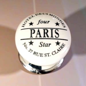 May include: A white ceramic cabinet knob with black text that reads "HOTEL DEUS MONDES four PARIS Star No. 37 RUE ST. CLAIRE". The knob has a silver metal base.
