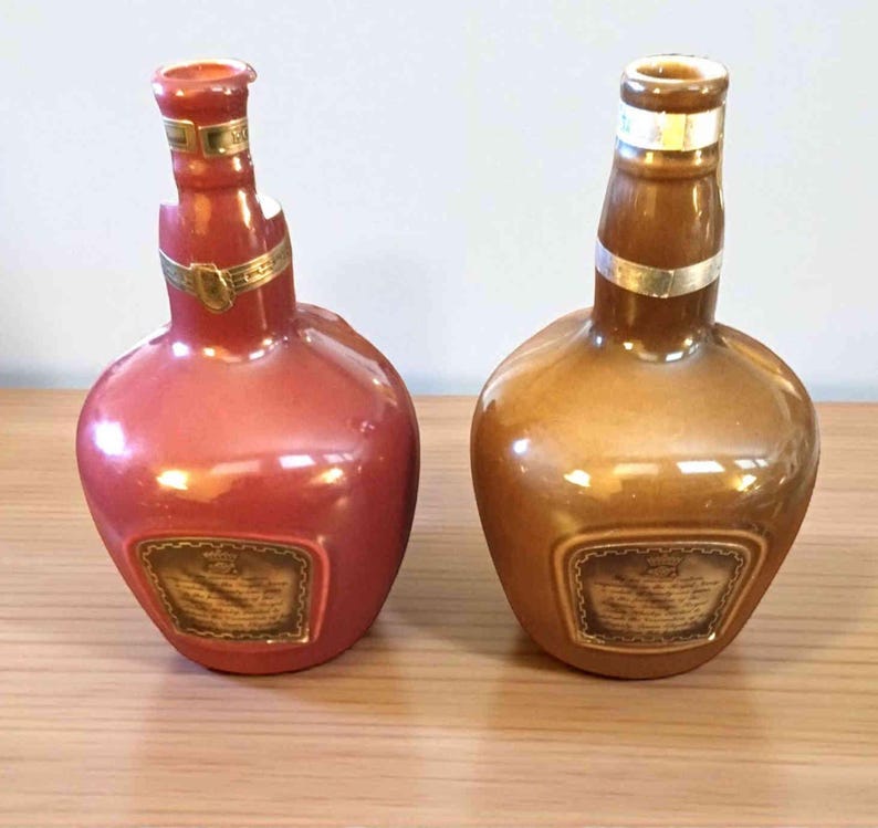 K&ouml;nnte beinhalten: Zwei dekorative Keramikflaschen, eine rot-rosa und die andere goldbraun. Beide haben einen abgerundeten Boden, einen schmalen Hals und ein goldenes Band. Jede Flasche hat ein rechteckiges Etikett mit Text.