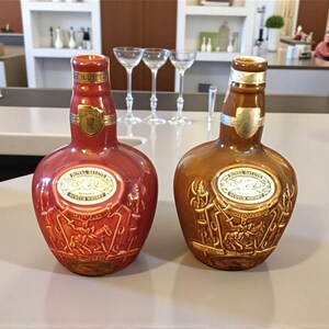K&ouml;nnte beinhalten: Zwei dekorative Lik&ouml;rflaschen, eine rote und eine braune, mit goldenen Akzenten und gepr&auml;gten Designs. Jede Flasche hat ein goldenes Etikett mit Text und einem Schildemblem. Die Flaschen stehen auf einer hellen Oberfl&auml;che.