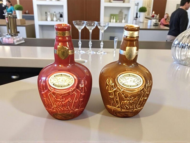 K&ouml;nnte beinhalten: Zwei dekorative Lik&ouml;rflaschen, eine rote und eine braune, mit goldenen Akzenten und gepr&auml;gten Designs. Jede Flasche hat ein goldenes Etikett mit Text und einem Schildemblem. Die Flaschen stehen auf einer hellen Oberfl&auml;che.