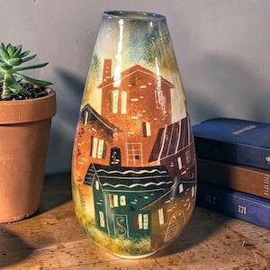 May include: A decorative, hand-painted ceramic vase with a unique village scene. The vase features a tall, slender shape with a wide base and a narrow opening. The artwork depicts houses in shades of brown, green, and white, with a gradient background.