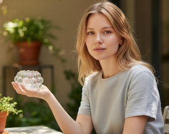May include: A clear, round glass candle holder with a bubble-like design. The candle holder is held in a person's hand. The background includes greenery and a wooden table.