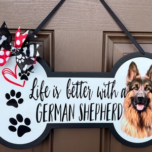 May include: Bone-shaped wooden sign with the text "Life is better with a GERMAN SHEPHERD." Features a German Shepherd portrait, paw prints, and a red heart. A decorative bow with black and white patterns and red polka dots is attached.