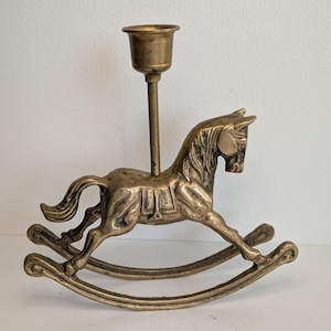 May include: A brass candlestick holder shaped like a rocking horse. The horse figure features a detailed mane, tail, and saddle, with a vertical rod extending from its back to support a cylindrical candle cup at the top, set on curved rockers.