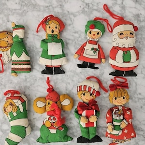 May include: A selection of vintage Christmas tree decorations. The fabric ornaments depict festive figures such as Santa, elves, and angels. Each decoration has a loop for hanging. The colour scheme includes red, green, white, and yellow.