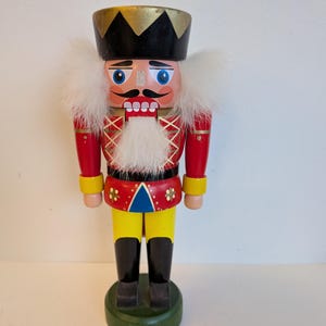 Peut inclure: Figurine de casse-noisette en bois. La figurine porte un chapeau noir avec des bordures dorées, une veste rouge avec des accents jaunes et un pantalon jaune. Il a une barbe blanche et une expression sévère. Le casse-noisette repose sur une base circulaire verte.