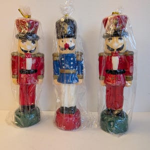 May include: Three decorative nutcracker figurines in clear plastic wrap. Two are red with black hats and gold accents, and one is blue with a black hat. Each has a painted face and stands on a green or red base.