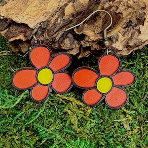 Puede incluir: Un par de pendientes con forma de flor. Cada pendiente presenta una flor roja con centro amarillo y contorno negro. Los pendientes están colocados sobre un fondo de musgo verde y un trozo de madera.