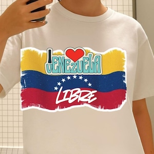 May include: White t-shirt featuring a graphic of the Venezuelan flag with the words "I Love Venezuela" and "Libre" in a stylized font. The flag design includes yellow, blue, and red stripes with white stars.