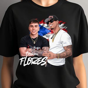 May include: Black t-shirt featuring a graphic of two men with tattoos, gold chains, and sunglasses, set against a Puerto Rican flag. The text "team FLORES" is displayed in white cursive font.