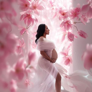 May include: A sheer, light pink maternity gown with an off-the-shoulder neckline and a flowing, floor-length skirt. The garment is draped in soft, translucent fabric that reveals the silhouette, surrounded by large, vibrant pink magnolia flowers.