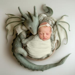 May include: A newborn baby girl sleeping peacefully in a woven basket, nestled next to a soft, green, felt dragon with wings. The baby is wearing a white swaddle and a flower headband.