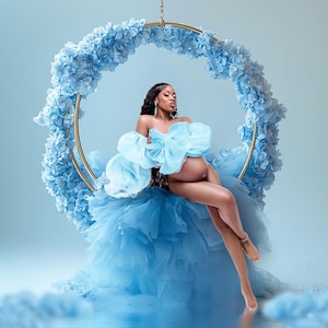 May include: A pregnant woman wearing a light blue tulle dress sits on a swing decorated with blue flowers. The swing is suspended from the ceiling against a light blue background.