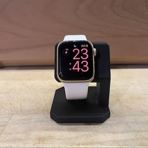 May include: A white Apple Watch Series 6 with a gold aluminium case is on a black charging stand. The watch face displays the time as 11:43 PM and the date as Fri 5. A lightning bolt icon indicates the watch is charging.