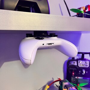 May include: A white Xbox controller is mounted upside down on a shelf with a black remote control on the shelf above it. There are several small drones and remote controls on the shelf below the controller.