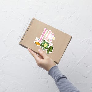 May include: A sticker with a cartoon frog holding a yellow object, and the text "Inverted Frog Energy" in pink and white. The frog is green and is on a green lily pad. Pink and yellow starbursts are in the background.