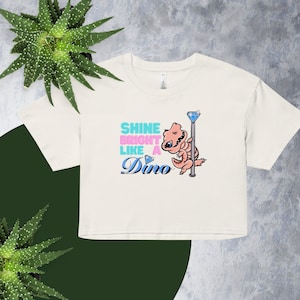 May include: Cream-colored crop top with the text "Shine Bright Like A Dino" in blue, pink, and blue script. A cartoon dinosaur is holding a pole with a diamond at the top. The shirt is on a green and gray background.