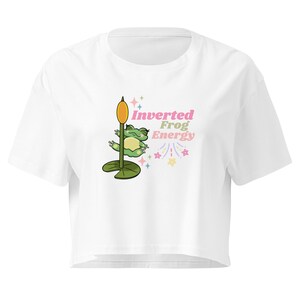 May include: A white cropped t-shirt featuring a graphic of a green frog hanging upside down from a cattail plant on a lily pad. The text Inverted Frog Energy is printed in pink and green, accompanied by small colourful star and sparkle icons.