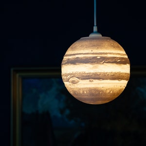 Może przedstawiać: Białe wiszące oświetlenie z okrągłym, kremowym kloszem przypominającym planetę Jowisz. Klosz ma brązowe i białe pasy oraz fakturowaną powierzchnię.