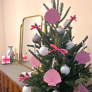 K&ouml;nnte beinhalten: Ein geschm&uuml;ckter Weihnachtsbaum mit rosa Muschelschmuck, wei&szlig;en und silbernen Kugeln und rosa Schleifen. Der Baum steht vor einem neutralen Hintergrund, mit einem Spiegel und Geschenken im Hintergrund.