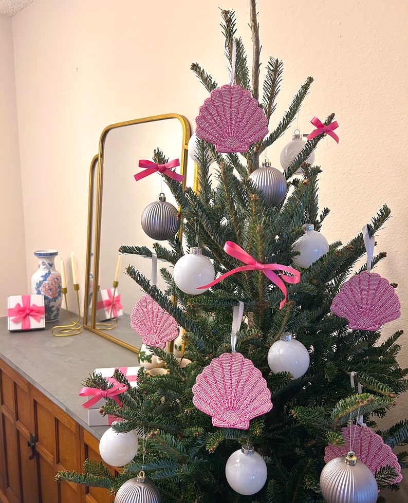 K&ouml;nnte beinhalten: Ein geschm&uuml;ckter Weihnachtsbaum mit rosa Muschelschmuck, wei&szlig;en und silbernen Kugeln und rosa Schleifen. Der Baum steht vor einem neutralen Hintergrund, mit einem Spiegel und Geschenken im Hintergrund.