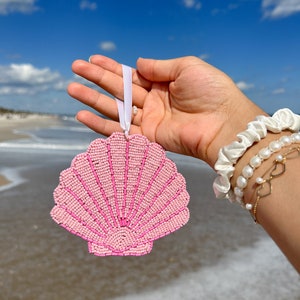 K&ouml;nnte beinhalten: Ein rosa, perlenbesetzter Muschelschmuck mit wei&szlig;em Band, vor blauem Himmel und Ozean. Der Schmuck hat eine F&auml;cherform mit detailliertem Perlendesign. Das Thema ist K&uuml;stendekoration.