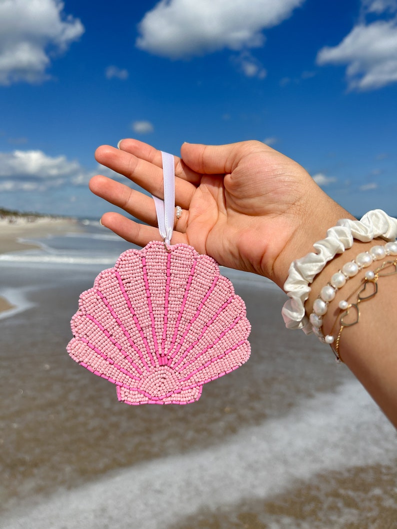 K&ouml;nnte beinhalten: Ein rosa, perlenbesetzter Muschelschmuck mit wei&szlig;em Band, vor blauem Himmel und Ozean. Der Schmuck hat eine F&auml;cherform mit detailliertem Perlendesign. Das Thema ist K&uuml;stendekoration.