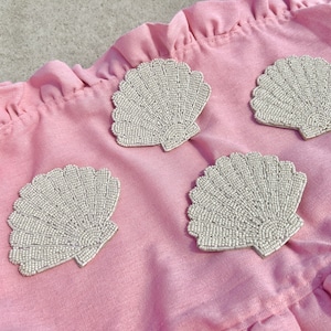 May include: Four seashell-shaped coasters with a beaded design in a neutral color. The coasters are arranged on a pink, ruffled fabric. The coasters are a decorative item for a coastal or beach-themed home.
