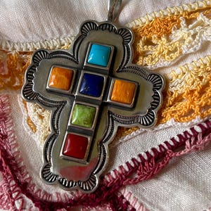May include: Sterling silver multi-stone cross pendant.  The cross features eight square-shaped stones in vibrant colors including turquoise, lapis lazuli, carnelian, and green.  The ornate silver frame has a southwestern-style design. The pendant is displayed on a crocheted fabric.