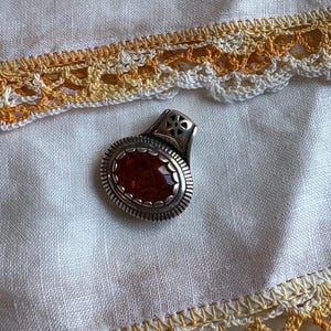 May include: A silver pendant with an oval, amber-colored stone. The pendant has a decorative border and a small cross design on the top. The pendant is set on a white cloth with a yellow and white lace trim.