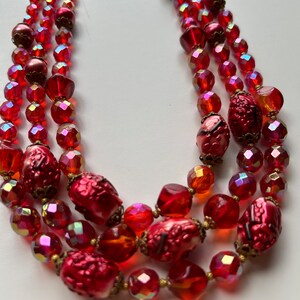May include: A multi-strand necklace made of red and orange glass beads. The necklace features a variety of bead shapes and sizes, including round, oval, and faceted beads. The necklace is finished with a gold-tone clasp.