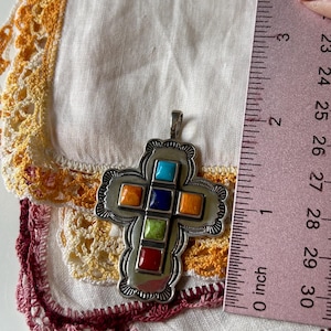 May include: A silver cross pendant with a detailed design and inlaid colorful stones. The stones are turquoise, orange, blue, green, and red. The pendant is about 1 inch in size.