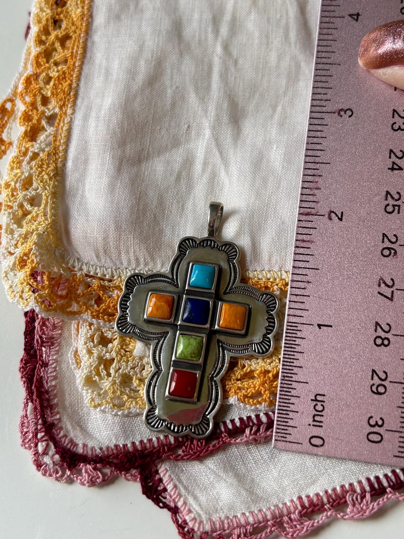 May include: A silver cross pendant with a detailed design and inlaid colorful stones. The stones are turquoise, orange, blue, green, and red. The pendant is about 1 inch in size.