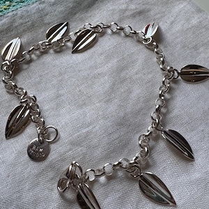 May include: A silver bracelet with leaf-shaped charms. The bracelet features a chain design with multiple leaf pendants and a small round tag. The leaves have a polished, reflective surface, and the bracelet is displayed on a white fabric.