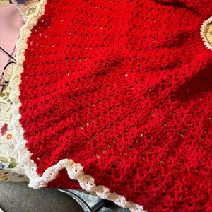May include: A circular, red crocheted tree skirt featuring a textured, open-stitch pattern throughout the main body. The outer edge is finished with a wide, scalloped white crocheted border that creates a decorative, wavy trim around the perimeter.