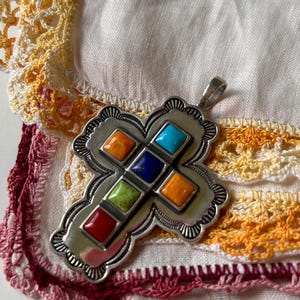 May include: A silver cross pendant with a decorative border and inlaid colorful stones. The stones are red, orange, yellow, green, blue, and turquoise.