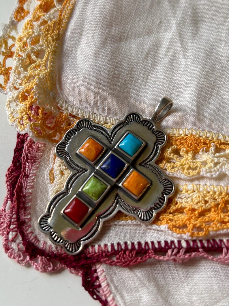 May include: A silver cross pendant with a decorative border and inlaid colorful stones. The stones are red, orange, yellow, green, blue, and turquoise.