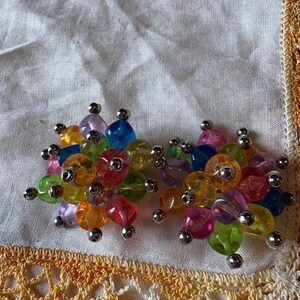 May include: A pair of colorful beaded earrings. Each earring features a cluster of translucent beads in various colors, including pink, blue, yellow, and green. The beads are connected with small silver-colored metal accents. The earrings are set against a white cloth with a yellow lace trim.