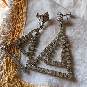 May include: A pair of silver-toned, rhinestone-studded earrings. The earrings feature a geometric design with multiple layered triangles. The earrings are set against a white cloth with a yellow lace trim.