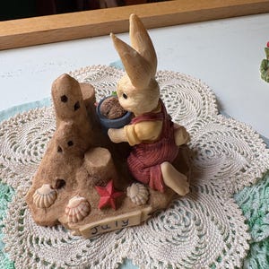 May include: A figurine of a bunny wearing overalls, holding a pot of soil, and sitting on a sandy beach scene. The base includes seashells, a red starfish, and the word "July".