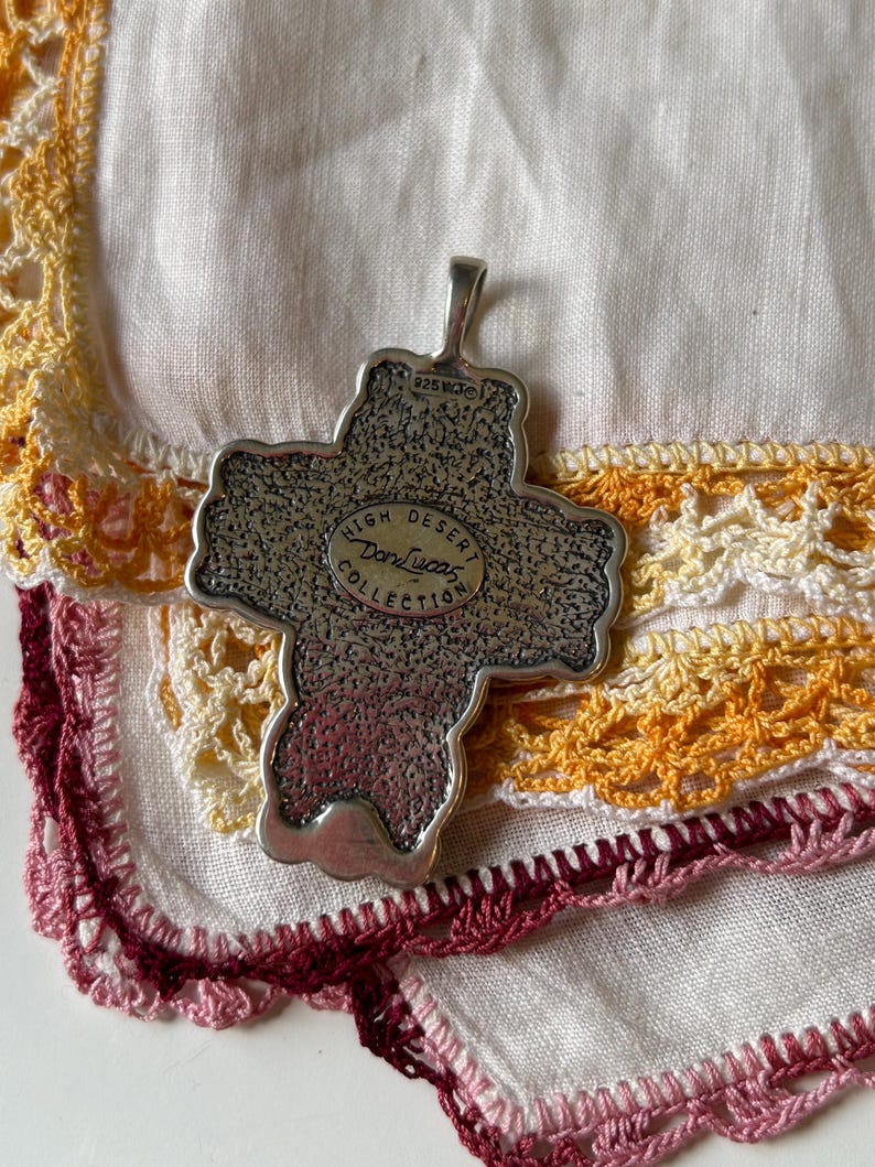 May include: Silver cross pendant with a textured surface and the words "High Desert Don Lucas Collection" inscribed on the front. The pendant has a loop at the top for attaching a chain.