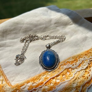 May include: A silver chain necklace with a blue gemstone pendant. The pendant is oval shaped and set in a silver frame with intricate details. The necklace is lying on a white fabric with a lace trim.