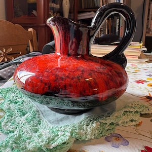 May include: A glossy, red and black ceramic pitcher with a large, curved black handle. The pitcher has a wide base and a narrow spout. It sits on a green crocheted doily. The pitcher is likely vintage or antique.