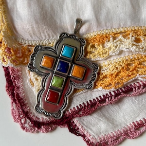 May include: A silver cross pendant with a detailed design and colorful stones. The stones are red, orange, yellow, green, blue, and turquoise.