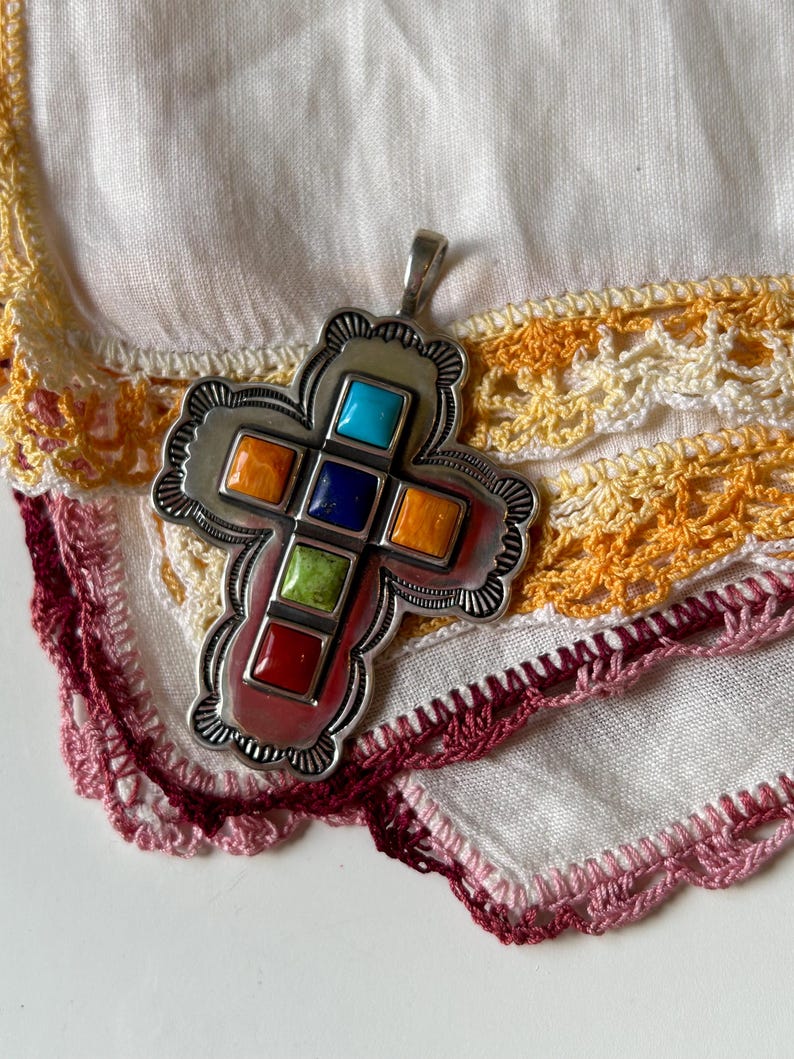 May include: A silver cross pendant with a detailed design and colorful stones. The stones are red, orange, yellow, green, blue, and turquoise.