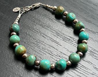 May include: A turquoise and silver beaded bracelet with a silver clasp. The bracelet is made with round turquoise beads and silver spacer beads.