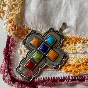 May include: A silver cross pendant with a southwestern design. The cross is decorated with eight colorful stones in a variety of colors, including red, orange, yellow, green, blue, and turquoise.
